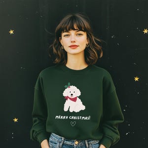 Op de afbeelding: Een donkergroene sweatshirt met een illustratie van een witte poedel met een rode bandana en hulst. De tekst "MERRY CHRISTMAS!" staat onder de hond, met een hartsymbool. De achtergrond is donker met gouden sterren.