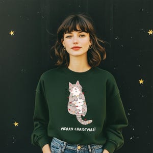 Op de afbeelding: Een donkergroene sweatshirt met een grijze kat met een glimlach, versierd met kerstverlichting. De tekst "MERRY CHRISTMAS!" staat eronder. De achtergrond is donker met gouden sterren.