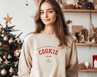 Pode incluir: Um moletom bege com o texto "PROUD MEMBER OF THE COOKIE LOVER CLUB" em vermelho e verde. O moletom é um modelo de gola redonda e está sendo usado por uma pessoa. O fundo inclui uma árvore de Natal e prateleiras decorativas.