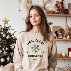 Op de afbeelding: Een beige sweatshirt met de tekst "MRS. CLAUS CHRISTMAS BAKERY Homemade Christmas Cookies" in groen en rood. Een sneeuwvlokkoekjesafbeelding staat in het midden. De sweatshirt wordt gedragen door een persoon voor een kerstboom.
