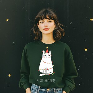 Op de afbeelding: Een donkergroene sweatshirt met een witte kattenprint met een kerstmuts en lichtsnoeren. De woorden "MERRY CHRISTMAS" staan eronder. De sweatshirt wordt gecombineerd met een blauwe jeans.
