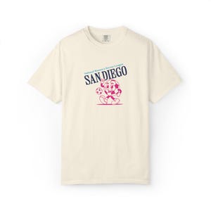 May include: Cream-colored t-shirt featuring a cartoon soccer ball character with a smiling face, holding a soccer ball. The text "National Women's Soccer League SAN DIEGO" is printed above the character in navy blue and pink.