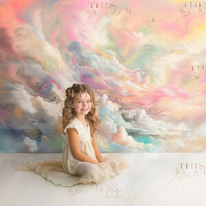 Puede incluir: Una niña pequeña con el pelo largo y rubio se sienta frente a un telón de fondo pintado de colores. El telón de fondo presenta un cielo con nubes en tonos de rosa, azul y amarillo. La niña lleva un vestido blanco y tiene una corona de flores en el pelo.