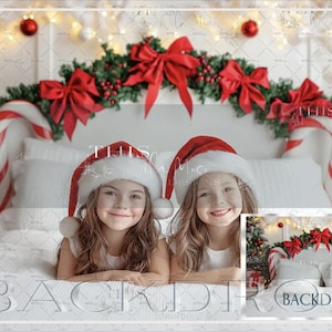 May include: Two girls wearing Santa hats are lying on a white bed with a candy cane headboard. The bed is decorated with a garland of red bows and greenery. The background is a white wall with a Christmas tree and presents.