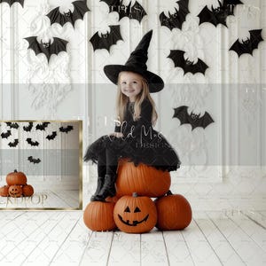 May include: A young girl dressed as a witch sits on a stack of orange pumpkins in front of a white wall with black bats hanging from the ceiling. The text "BACKDROP" is visible on a framed picture behind the pumpkins.