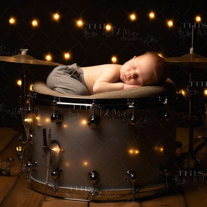 May include: A newborn baby sleeping on a drum with a cymbal behind it. The drum is decorated with a white cushion and the baby is wearing grey pants. The background is dark with a string of lights.