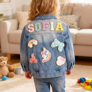 Könnte beinhalten: Hellblaue Jeansjacke mit dem Namen "SOFIA" in bunten, gestickten Buchstaben auf dem Rücken. Die Jacke ist mit Aufnähern von Regenbogen, Einhorn und Wolken verziert. Das Kind trägt eine passende blaue Jeans.