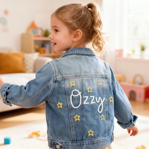 May include: A light blue denim jacket with the name "Ozzy" embroidered in white script on the back, surrounded by yellow star accents. The jacket is a classic style, suitable for children's wear.