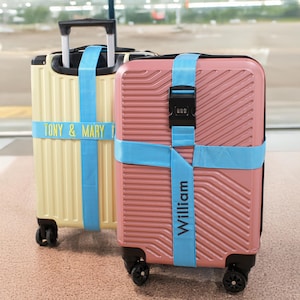May include: Two rolling suitcases, one pale yellow and one pink, secured with light blue straps. The yellow suitcase has the text "TONY & MARY" and the pink suitcase has the text "William". Both have black wheels and handles.