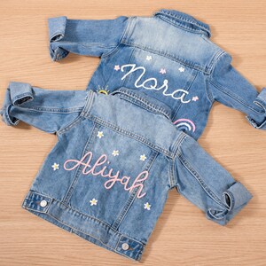May include: Two light blue denim jackets with rolled-up sleeves. One jacket has the name "Nora" embroidered in white with a rainbow and sun design. The other jacket has the name "Aliyah" embroidered in pink with flower accents.