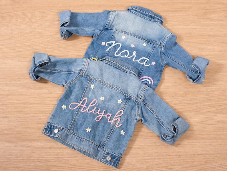 May include: Two light blue denim jackets with rolled-up sleeves. One jacket has the name "Nora" embroidered in white with a rainbow and sun design. The other jacket has the name "Aliyah" embroidered in pink with flower accents.