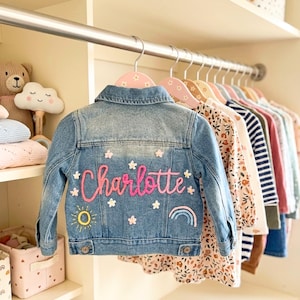 May include: A light blue denim jacket embroidered with the name "Charlotte" in pink, featuring flower and star motifs, a sun, and a rainbow. The jacket hangs on a pink floral hanger in a wardrobe with other children's clothes.