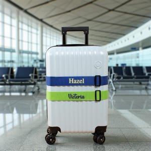 Könnte beinhalten: Weißer Rollkoffer mit blauen und grünen Gurten. Der blaue Gurt trägt den Namen "Hazel" in goldfarbenen Buchstaben, und der grüne Gurt trägt "Victoria". Der Koffer hat einen ausziehbaren Griff und vier Räder.