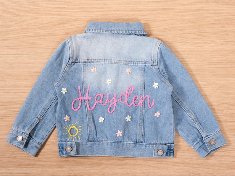 May include: Light blue denim jacket with the name "Hayden" embroidered in pink script on the back. The jacket is decorated with embroidered flowers and a yellow sun. A classic, stylish piece.