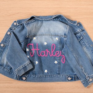 May include: Light blue denim jacket with the name "Harley" embroidered in pink script on the back. White flower accents are scattered around the name. The jacket has button closures and pockets.