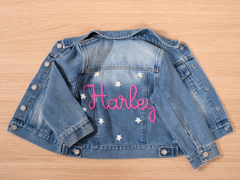 May include: Light blue denim jacket with the name "Harley" embroidered in pink script on the back. White flower accents are scattered around the name. The jacket has button closures and pockets.