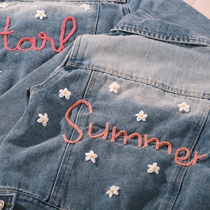 May include: Two denim jackets with light blue wash. One jacket has the name "Harli" embroidered in pink yarn, the other has "Summer". Both jackets are decorated with small white daisy appliqu&eacute;s.