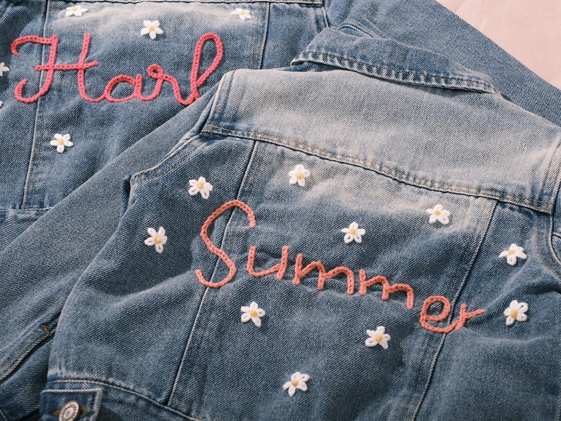 May include: Two denim jackets with light blue wash. One jacket has the name "Harli" embroidered in pink yarn, the other has "Summer". Both jackets are decorated with small white daisy appliqu&eacute;s.