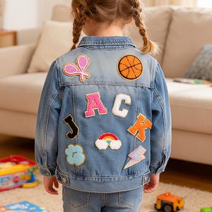 Puede incluir: Chaqueta vaquera azul claro con parches coloridos, incluyendo una pelota de baloncesto, un arcoíris y un rayo. La chaqueta tiene las letras "JAC K" aplicadas en la espalda. El niño lleva vaqueros azules.