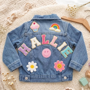 May include: A blue denim jacket featuring the name HALLIE in large, colorful, textured fabric letters across the back. The jacket is decorated with embroidered patches including a cupcake, a rainbow, a pink lightning bolt smiley face, and two flowers.
