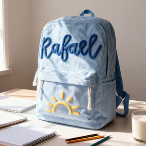 May include: Pale blue corduroy backpack with the name "Rafael" embroidered in blue script. Features a front pocket with a yellow and white sun design. Includes adjustable straps and a top handle. Ideal for school or travel.