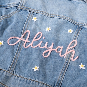 May include: Light blue denim jacket with the name "Aliyah" embroidered in pink script. The jacket is decorated with small white daisy appliqués. A classic children's denim jacket.