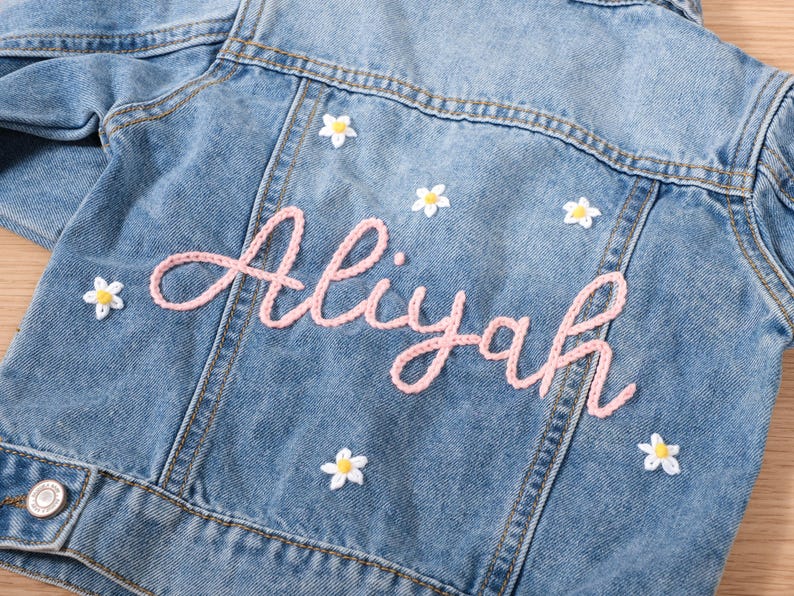 May include: Light blue denim jacket with the name "Aliyah" in pink script. The jacket is decorated with small white and yellow daisy appliqu&eacute;s. A classic children's denim jacket.