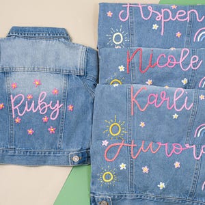 May include: Pale blue denim jackets with embroidered names, flowers, suns, and rainbows. The jackets are personalised with names such as "Ruby", "Aspen", "Nicole", "Karli", and "Aurora". The jackets are decorated with pink and yellow embroidery.