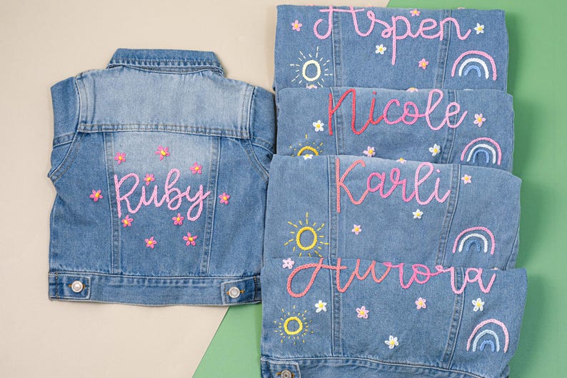May include: Pale blue denim jackets with embroidered names, flowers, suns, and rainbows. The jackets are personalised with names such as "Ruby", "Aspen", "Nicole", "Karli", and "Aurora". The jackets are decorated with pink and yellow embroidery.
