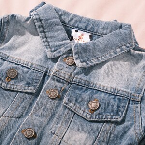 May include: Close-up of a light blue denim jacket with a classic collar and two chest pockets. The jacket features silver-colored metal buttons and a faded wash effect, giving it a vintage look. The collar lining has a white and black polka dot pattern.