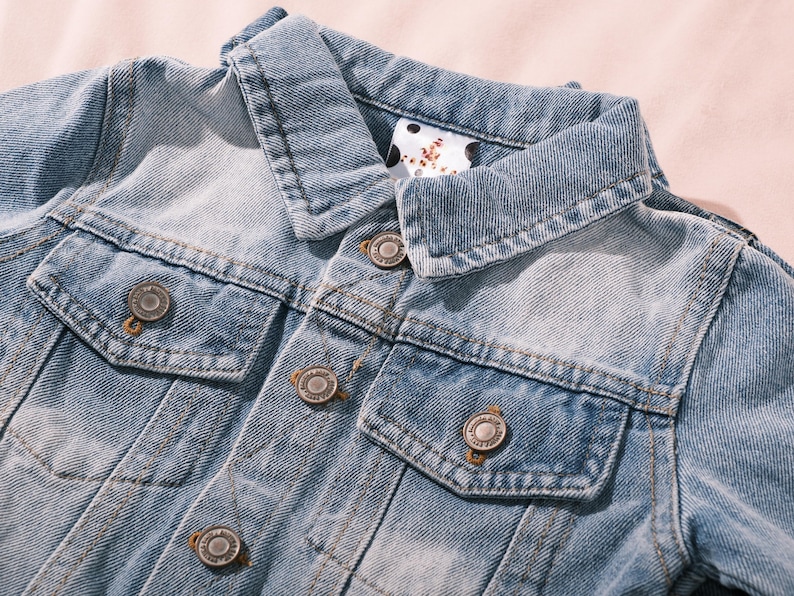 May include: Close-up of a light blue denim jacket with a classic collar and two chest pockets. The jacket features silver-colored metal buttons and a faded wash effect, giving it a vintage look. The collar lining has a white and black polka dot pattern.