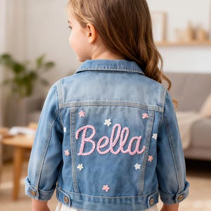 May include: A light blue denim jacket with the name "Bella" embroidered in pink script on the back, accented with small pink and white flower appliqu&eacute;s. The jacket has button closures and classic denim details.