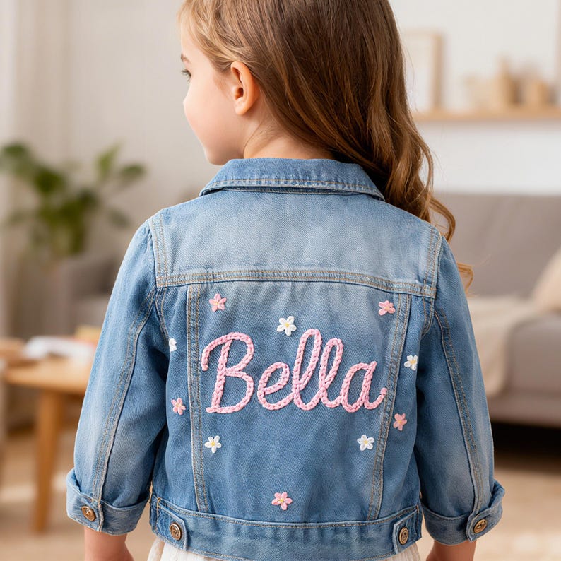 May include: A light blue denim jacket with the name "Bella" embroidered in pink script on the back, accented with small pink and white flower appliqu&eacute;s. The jacket has button closures and classic denim details.