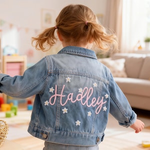 May include: A light blue denim jacket with the name "Hadley" embroidered in pink script on the back, surrounded by white flower accents. The jacket has a classic collar and button closures, suitable for children.