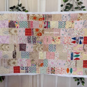 May include: A patchwork quilt with a white border. The quilt is made of various fabric squares in shades of pink, beige, and red, with floral and geometric patterns. The text "Desi company" is visible on the quilt.