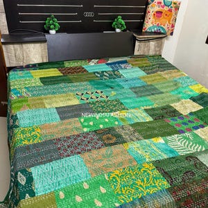 Könnte beinhalten: Eine Patchwork-Steppdecke in Grün-, Türkis- und Olivtönen bedeckt ein Bett. Die Steppdecke besteht aus verschiedenen Stoffquadraten mit unterschiedlichen Mustern und Texturen. Das Kopfteil ist schwarz mit silbernen Akzenten und kleinen grünen Pflanzen auf jeder Seite.