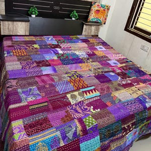 May include: A patchwork quilt in shades of purple, pink, and blue covers a bed. The quilt is made of various fabric squares stitched together. The text "NEW INDIA FASHION" is visible on the quilt. A decorative pillow with an elephant design rests on the bed.