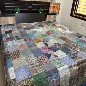 May include: A patchwork quilt covers a bed, featuring a variety of fabrics in muted tones of blue, green, and brown. The headboard is dark with silver accents. A decorative pillow with a colorful pattern rests on the bed.