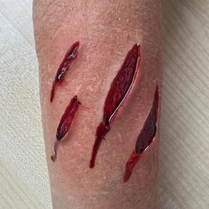 Pode incluir: Close-up de um braço com quatro lacerações profundas e sangrentas. Os cortes são vermelhos e cheios de sangue, parecendo frescos. A pele é pálida e texturizada.