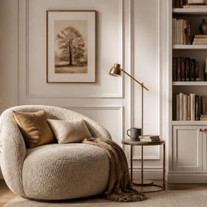May include: A round, cream-colored bouclé armchair featuring a curved back, accompanied by a brown throw pillow, a cream pillow, and a brown throw blanket. Beside the chair sits a small round metal side table with a lamp, a mug, and books.