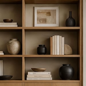 Puede incluir: Una estantería de madera alta con varios niveles que contiene diversos objetos decorativos, incluyendo jarrones de cerámica en tonos gris, negro y beige, pilas de libros con portadas neutras, cuencos de madera y cuadros enmarcados.