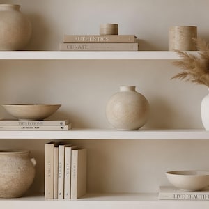 Könnte beinhalten: Ein weißes Regal mit beigen Keramikvasen, Schalen und Büchern. Die Bücher tragen Titel wie Authentics, Curate, This is Home, Kinfolk und Live Beautiful. Einige Vasen enthalten getrocknetes Pampasgras, während andere leer sind.
