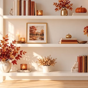Puede incluir: Tres estantes flotantes blancos dispuestos en una pared, decorados con artículos de temática otoñal que incluyen libros con lomos rojos y marrones, calabazas naranjas, jarrones con follaje, arte paisajístico enmarcado y velas de cristal.