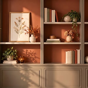 Puede incluir: Una estantería empotrada con pared trasera de color terracota y molduras grises, con múltiples estantes llenos de libros, plantas verdes en macetas, una lámina botánica enmarcada, pequeños jarrones y velas bajo luz natural.