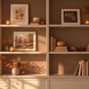 Puede incluir: Una estantería blanca empotrada con decoración otoñal, que incluye láminas botánicas enmarcadas, arte paisajístico, pilas de libros en tonos neutros, pequeñas calabazas decorativas, portavelas de cristal y jarrones con follaje naranja.