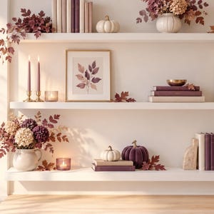 May include: Two white floating shelves decorated with autumn-themed items including small white and purple pumpkins, stacks of books, framed botanical art, dried flower arrangements in vases, lit candles in glass holders, and brass candlesticks.