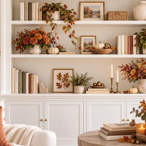 May include: A white built-in shelving unit decorated with autumn-themed items including orange and brown faux foliage, white pumpkins, assorted books, framed landscape art, brass candlesticks, and ceramic vases filled with seasonal floral arrangements.