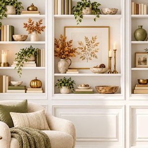 May include: A white built-in bookshelf filled with books, potted plants, decorative bowls, candles, and pumpkins. A cream-coloured armchair with a green throw blanket and pillows sits in front of the shelves, next to a small table with a plant.