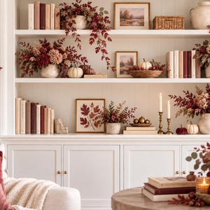 Op de afbeelding: Een witte inbouwkast versierd met herfstthema-items, waaronder boeken, ingelijste landschapskunst, keramische vazen gevuld met rode en crèmekleurige kunstbloemen, kleine witte pompoenen en koperen kandelaars met witte dinerkaarsen.