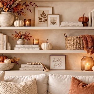 May include: A three-tiered shelving unit styled with autumn decor including white and orange pumpkins, dried foliage, candles, woven baskets, books with titles like A Kinder Home and Hello Fall, and framed botanical prints arranged on white shelves.